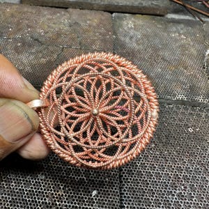 May include: A copper-colored pendant with a detailed, circular design. The pendant features an intricate pattern of woven wire, creating a floral or mandala-like shape. The piece is held against a textured metal surface.