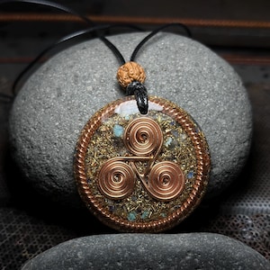 Peut inclure: Un pendentif circulaire présentant un symbole triskèle en cuivre composé de trois spirales, entouré de paillettes métalliques dorées et de petits fragments de pierre bleue, enchâssé dans un disque en résine avec un bord en fil de cuivre torsadé.
