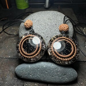 May include: Two round pendants with black centers and copper wire spirals. Each pendant has a brown bead at the top and hangs from a black cord. The pendants are displayed on a gray stone, suggesting a natural or spiritual theme.