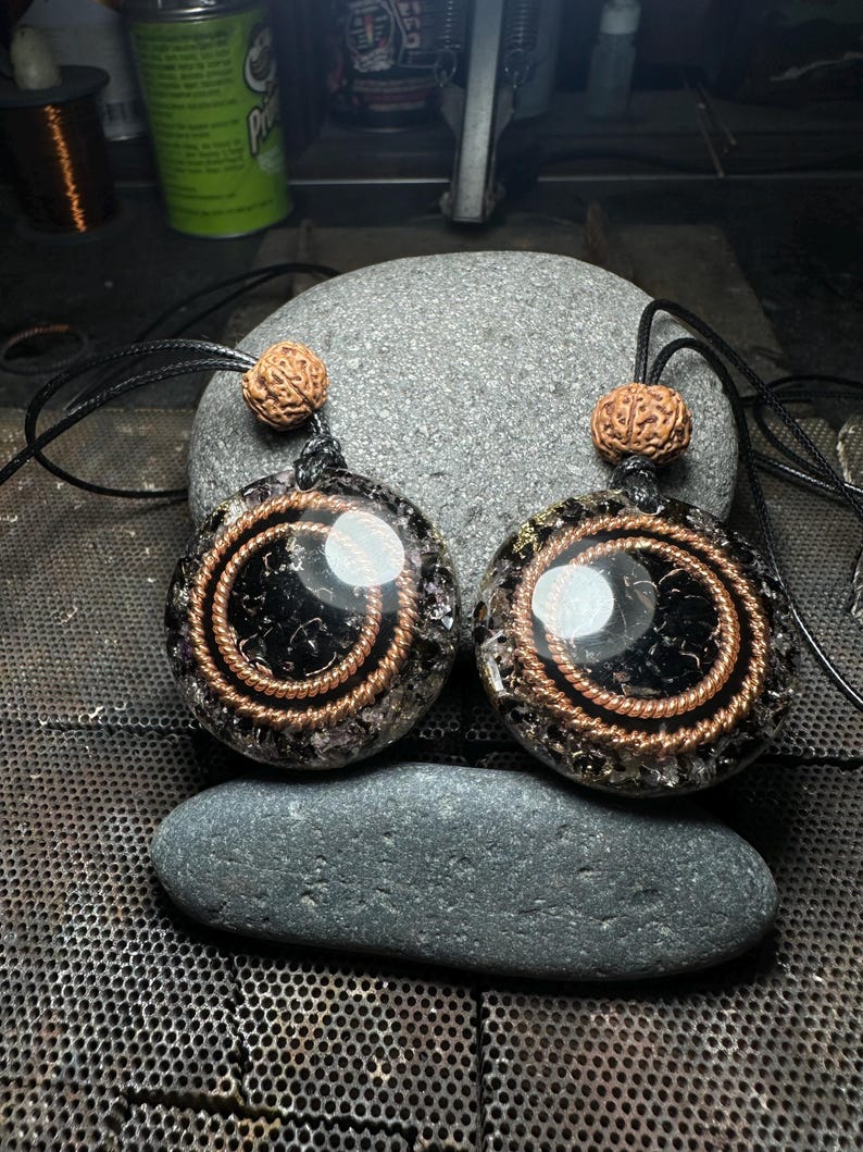 May include: Two round pendants with black centers and copper wire spirals. Each pendant has a brown bead at the top and hangs from a black cord. The pendants are displayed on a gray stone, suggesting a natural or spiritual theme.