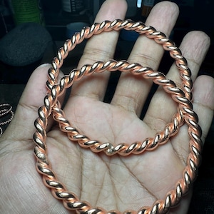 May include: Two copper-colored, twisted metal bangles. The bangles are circular and appear to be made of a series of intertwined strands. The metal has a shiny, reflective surface. The bangles are held in a person's hand.