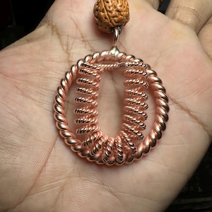 May include: A copper wire pendant featuring a circular frame made of twisted wire with two internal coiled wire springs. The pendant is suspended from a black cord and accented with a single textured brown seed bead at the top bail connection.