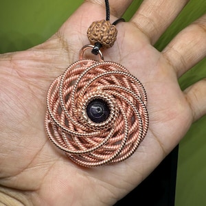 Può includere: Un ciondolo a spirale in filo di rame con una pietra viola scuro al centro. Il ciondolo è attaccato a un cordoncino nero con una perla di legno marrone.
