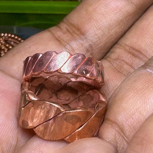 May include: Two copper-colored rings with a braided design. The rings are made of a shiny, metallic material and have a textured surface. The rings are displayed in a hand.