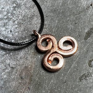 Op de afbeelding: Een koperkleurige triskelion hanger met drie in elkaar grijpende spiraalarmen en een getextureerde, metalen afwerking. De hanger hangt aan een dunne, zwarte koordketting en toont een opvallend cirkelvormig ontwerp met een centrale lus.