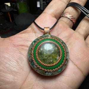 Peut inclure: Un pendentif circulaire avec un cœur en résine moucheté vert et or, entouré d'un anneau en fil de cuivre torsadé et d'une bande verte lisse, le tout dans un dôme en résine transparente avec des fragments minéraux et un cordon noir.