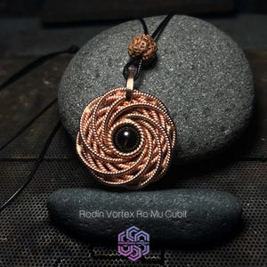 Può includere: Un ciondolo in rame Rodin Vortex Ra-Mu Cubit con una pietra nera al centro, appeso a un cordoncino nero con una perla marrone. Il ciondolo è appoggiato su una roccia grigia.