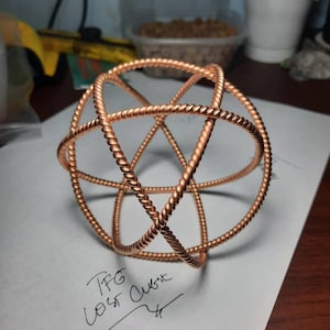 May include: A copper-toned, spherical sculpture with a woven, twisted wire design. The geometric form is made of intersecting rings, creating an open, three-dimensional shape. The sculpture rests on a white surface with handwritten text that reads "TFG Lost Cube".