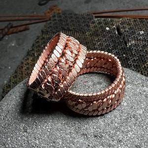 May include: Close-up of two copper-colored rings. The rings are made of small, interconnected, textured metal pieces. One ring is upright, while the other is lying on its side. The rings are resting on a gray, textured stone.