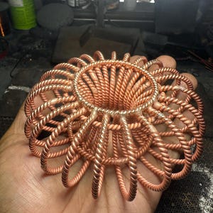 May include: A circular sculpture crafted from twisted copper wire, featuring a central ring connected to numerous radiating, arched wire loops. The metallic structure displays a warm, reddish-brown hue with visible solder points where the individual wire segments meet the central circular frame.
