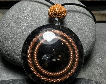 May include: A circular black resin pendant featuring two concentric copper wire coils embedded within the material. A small white triangular accent is visible near the top. The pendant hangs from a black cord adorned with a single textured brown bead.