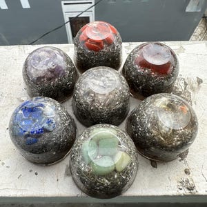 May include: Seven clear resin spheres, each containing different colored stones and crystals. The spheres are arranged in a circular pattern, with colors including red, purple, blue, green, and clear. The spheres are on a gray surface.