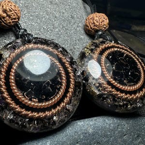 May include: Two pendant necklaces with black and copper spiral designs. Each pendant has a central white stone and is strung on a black cord. The top of each pendant features a brown, textured bead.