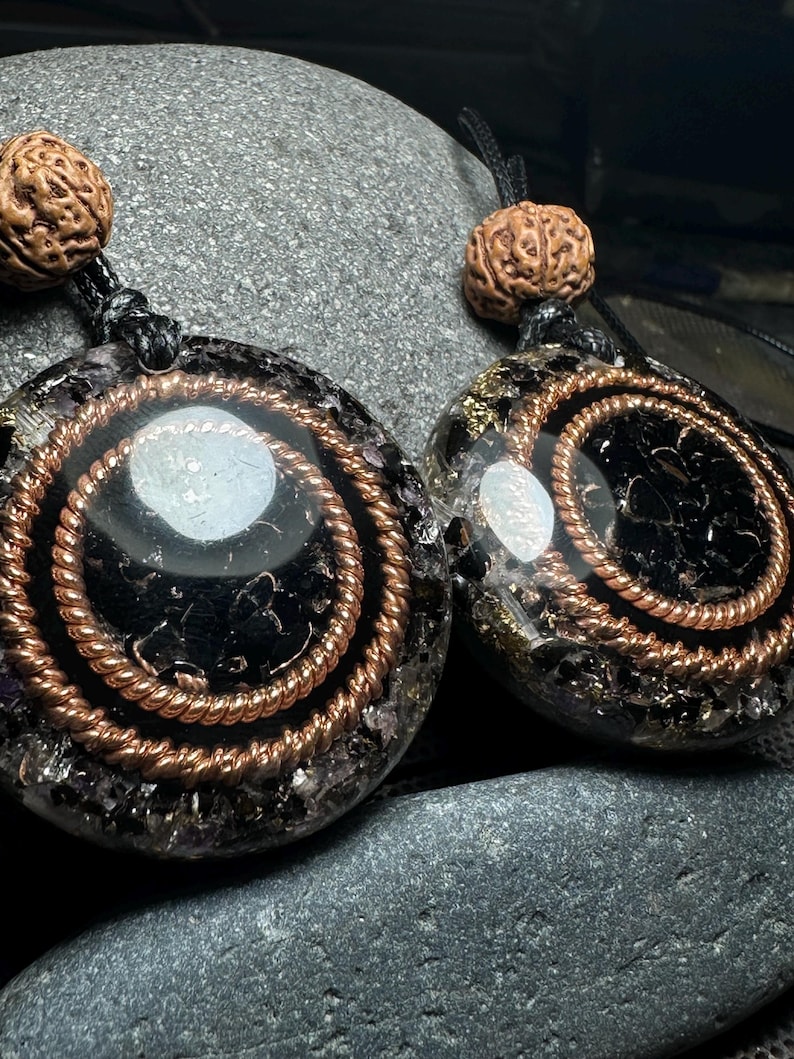 May include: Two pendant necklaces with black and copper spiral designs. Each pendant has a central white stone and is strung on a black cord. The top of each pendant features a brown, textured bead.