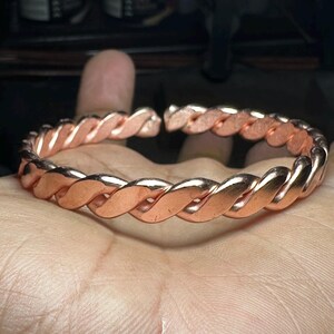 Könnte beinhalten: Ein kupferfarbenes, geflochtenes Armband. Das Armband hat ein gedrehtes Seildesign und ist an den Enden offen. Das Metall hat eine polierte Oberfläche, die das Licht reflektiert. Das Armband wird auf der Hand einer Person gezeigt.