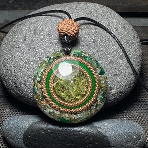Op de afbeelding: Een ronde hangerketting met een zwart koord. De hanger heeft een centrale groene steen, een koperdraadspiraal en een groene rand. De hanger is gevuld met groene en heldere stenen. Bovenop zit een bruine, getextureerde kraal.