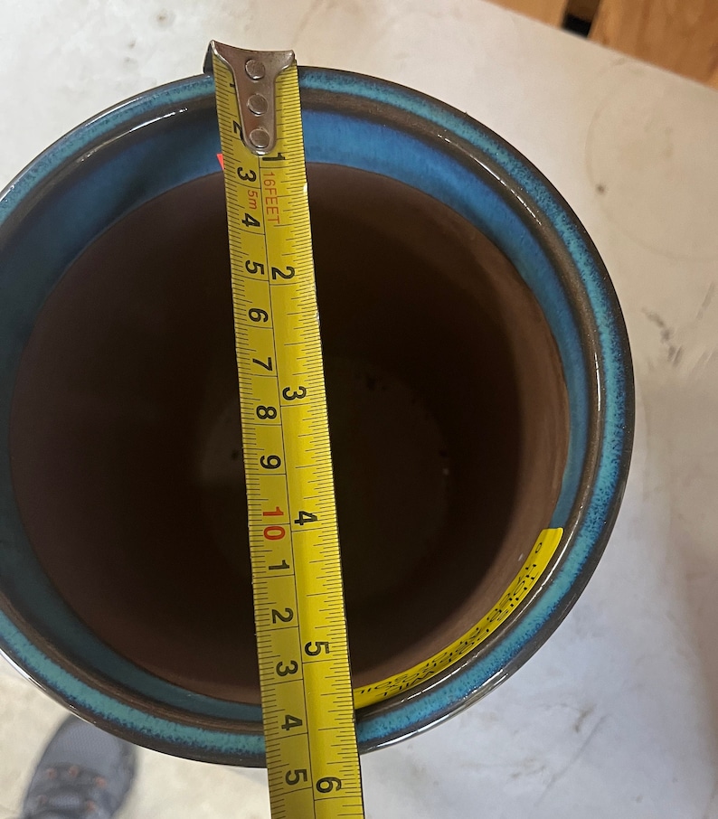 Op de afbeelding: Een blauwe en bruine keramische plantenpot met een gele meetlint ernaast. De tape meet 12,7 cm.