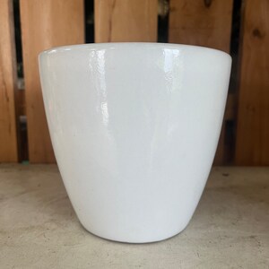 Op de afbeelding: Een witte keramische plantenpot met een gladde, glanzende afwerking. De pot heeft een afgeronde vorm en een brede opening.