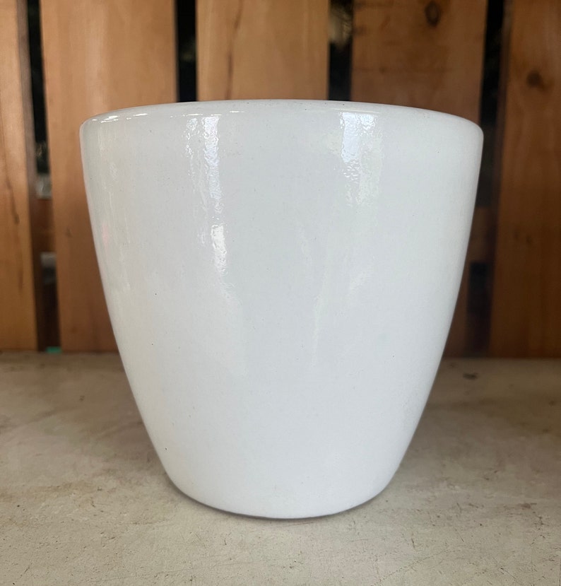 Op de afbeelding: Een witte keramische plantenpot met een gladde, glanzende afwerking. De pot heeft een afgeronde vorm en een brede opening.