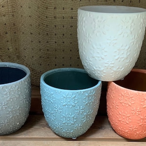 May include: Four ceramic flower pots in different colors: light blue, teal, white, and coral. Each pot has a raised floral pattern.