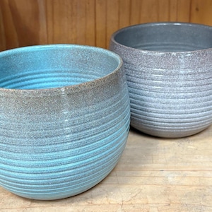 May include: Two ceramic planters with a textured, ribbed design. The planter on the left is a light blue color with a darker blue rim. The planter on the right is a light gray color with a darker gray rim.