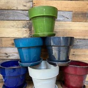 May include: Assortment of six glazed ceramic flower pots in various colors, including green, teal, gray, blue, white, and red. The pots are stacked in a pyramid shape.