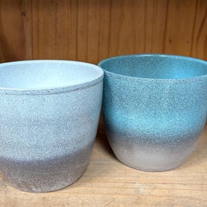 May include: Two ceramic planters with a speckled finish. The planter on the left is gray with a white gradient, while the planter on the right is blue with a white gradient.