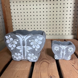 May include: Two gray concrete butterfly-shaped planters with white floral designs. The larger planter is in the foreground and the smaller planter is in the background.