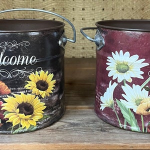May include: Two metal containers with handles, decorated with a floral pattern and the word "Welcome". The containers are brown and burgundy with sunflowers, daisies, and poppies.