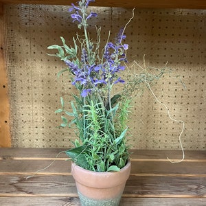 May include: A potted arrangement of faux lavender and greenery in a terracotta pot with a green glaze. The pot is sitting on a wooden surface.