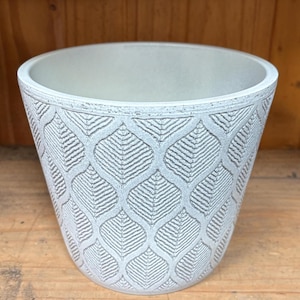 May include: A white ceramic planter with a leaf pattern. The planter has a slightly flared rim and a smooth, matte finish.