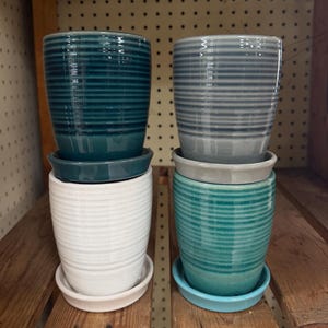 May include: Four cylindrical ceramic plant pots with horizontal ribbed textures and matching saucers. The pots are arranged in two stacks, featuring glossy finishes in deep teal, light gray, solid white, and a vibrant turquoise blue color.