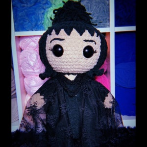 K&ouml;nnte beinhalten: Eine schwarze H&auml;kelpuppe mit einem schwarzen Spitzenkleid und einem schwarzen Dutt. Die Puppe hat gro&szlig;e schwarze Augen und ein rosafarbenes Gesicht.