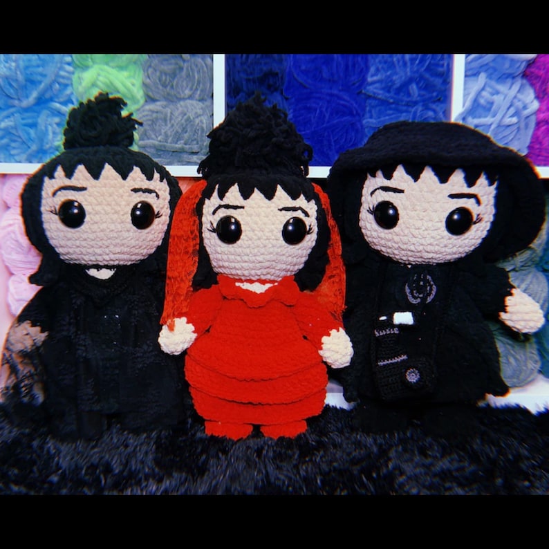 K&ouml;nnte beinhalten: Drei geh&auml;kelte Pl&uuml;schpuppen mit schwarzen Haaren und gro&szlig;en Augen. Die Puppen tragen verschiedene Outfits: eine tr&auml;gt ein schwarzes Kleid, eine tr&auml;gt ein rotes Kleid und eine tr&auml;gt einen schwarzen Mantel.