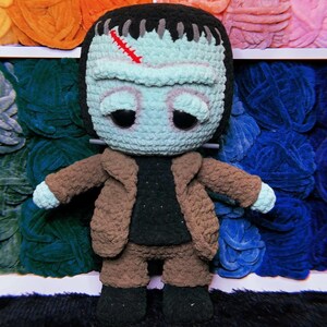 May include: A crocheted Frankenstein monster doll with a brown jacket, brown pants, and black shoes. The doll has a green face with a stitched scar and big, sad eyes.
