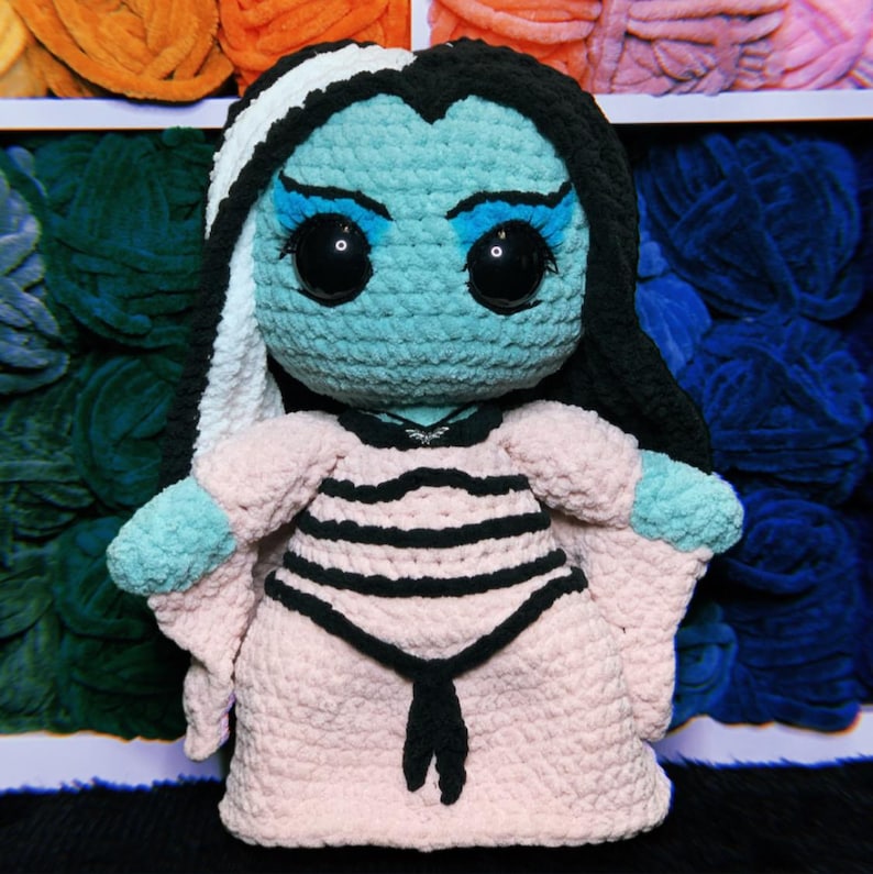 May include: A crocheted plush toy of a character with black and white hair, blue skin, and large eyes. The toy is wearing a pink dress with black stripes and a black bow.