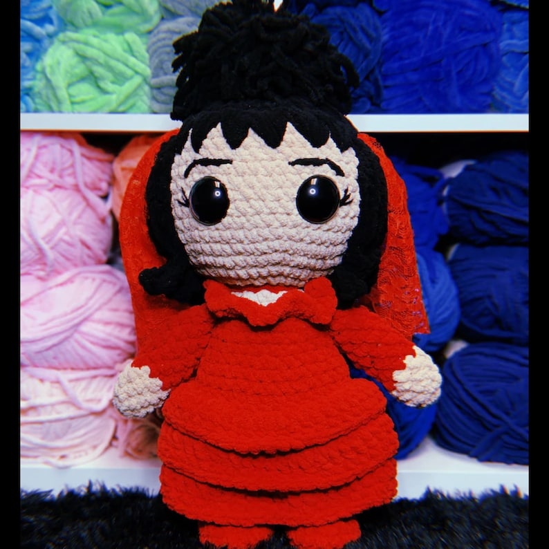 K&ouml;nnte beinhalten: Eine geh&auml;kelte Puppe mit schwarzen Haaren, einem roten Kleid und einem roten Schleier. Die Puppe hat gro&szlig;e schwarze Augen und ein wei&szlig;es Gesicht. Die Puppe sitzt auf einer flauschigen Oberfl&auml;che.