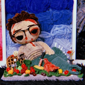 May include: A crocheted doll with brown hair and black-rimmed glasses, featuring red blood-like stains on the face and chest. The doll wears dark green pants and is surrounded by small crocheted fruits, a silver bucket, and a toy knife.
