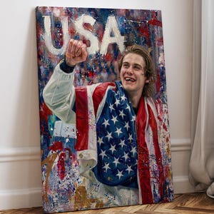 Pode incluir: Impress&atilde;o em tela com um retrato de uma pessoa sorrindo envolta em uma bandeira americana. A palavra "USA" est&aacute; pintada em letras brancas grandes no topo. A obra de arte tem um fundo colorido e abstrato com respingos de tinta.