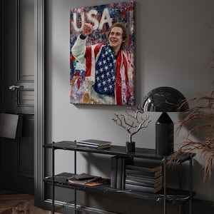 Pode incluir: Uma impress&atilde;o em tela emoldurada com uma pessoa envolta em uma bandeira americana, com a palavra "USA" vis&iacute;vel. A obra de arte &eacute; exibida acima de uma consola de m&aacute;rmore preto com objetos decorativos.