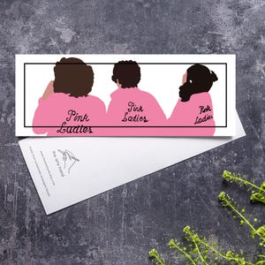 Può includere: Una carta rettangolare presenta tre figure con giacche rosa con la scritta "Pink Ladies" sulla schiena. Le figure hanno i capelli scuri e sono incorniciate in nero. La carta poggia su una superficie strutturata.
