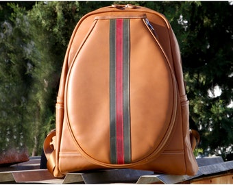 May include: A tan leather backpack featuring a large, oval-shaped front compartment with a central vertical stripe in dark green and red. The bag includes white contrast stitching, dual zipper pulls at the top, and side pockets on both sides.