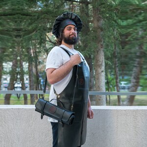 以下が含まれることがあります： 黒いシェフの帽子と赤いロゴのついたエプロンを着た人が、赤いロゴのついた黒い革製のナイフロールを持っています。