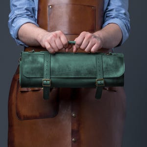 Puede incluir: Un estuche para cuchillos de cuero verde con hebillas y correas de latón. El estuche lo sostiene una persona que lleva un delantal de cuero marrón. El estuche para cuchillos tiene un asa y un cierre de solapa.