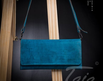 May include: A teal blue leather crossbody bag with a flap closure and a long strap. The bag is made in Georgia and has the word "Taia" written on the bottom right corner.