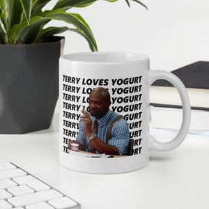 以下が含まれることがあります： 白地に黒と白の男性の画像と「TERRY LOVES YOGURT」というテキストが複数回繰り返されている白いセラミックマグカップ