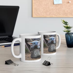 Puede incluir: Dos tazas de cer&aacute;mica blanca con un gr&aacute;fico de un personaje de piel azul sosteniendo una taza. El personaje lleva una t&uacute;nica marr&oacute;n y est&aacute; de pie en un entorno de bosque.
