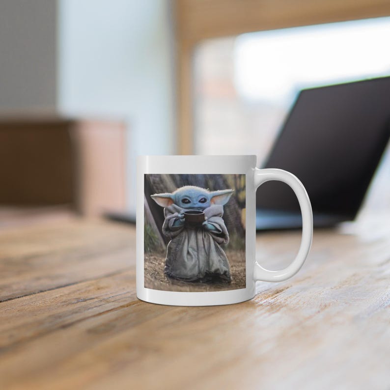 Puede incluir: Taza de cer&aacute;mica blanca con una imagen colorida de un personaje ficticio sosteniendo una taza. El personaje es azul, verde y marr&oacute;n con orejas grandes. La taza tiene un asa en el lado derecho.