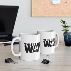 May include: Two white ceramic mugs with black text and a silhouette of a character from the Star Wars franchise. The text on the mugs reads "This is the Way".