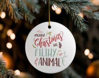 Op de afbeelding: Een witte, ronde kerstbal met de tekst "Merry Christmas ya Filthy Animal" in rode, groene en blauwe letters. De ornament hangt aan een gouden draad tegen een vage achtergrond van kerstboomtakken en lichtjes.